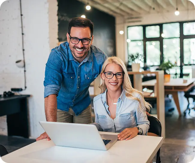 A man and a woman looking at the camera while working on a laptop